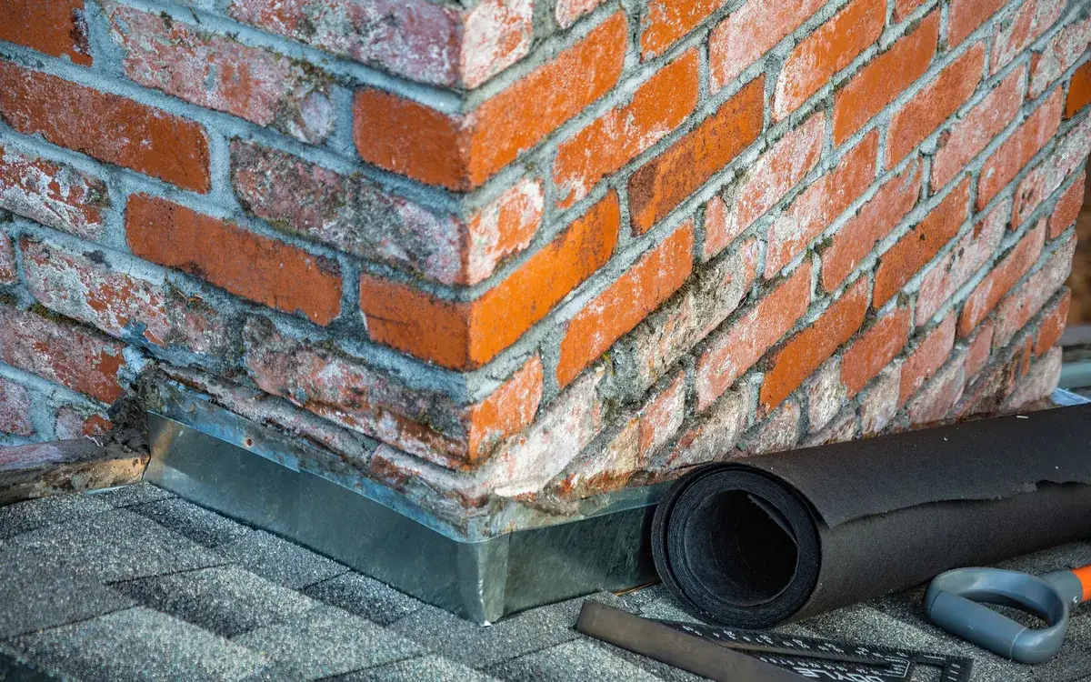 Metal flashing where a brick chimney meets the roof, next to a roll of underlayment