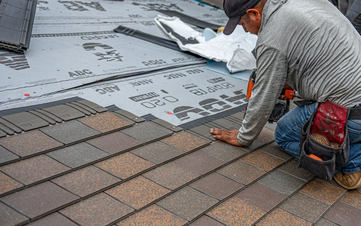Roofer kneeling on a roof while installing shingles over underlayment