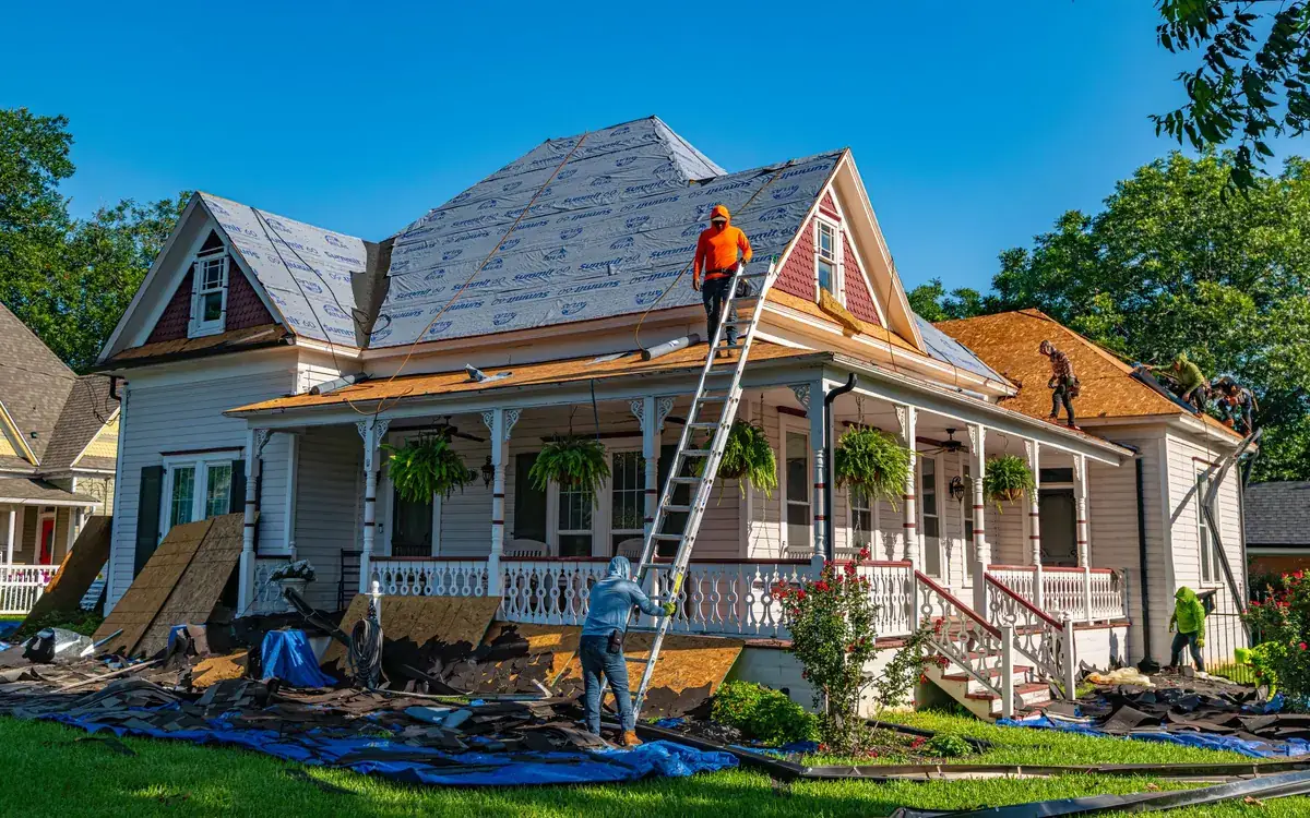 Roofers working on a two-story house with ladders and tarps around it