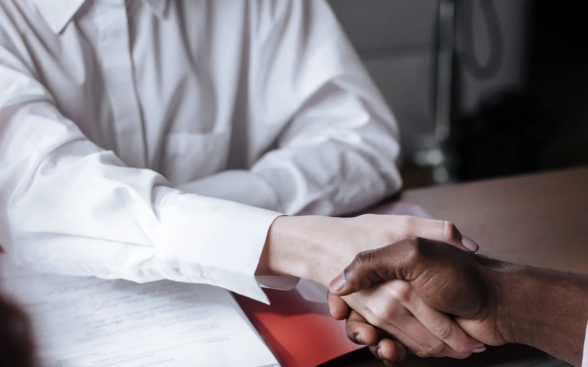Two people shaking hands across a desk with paperwork