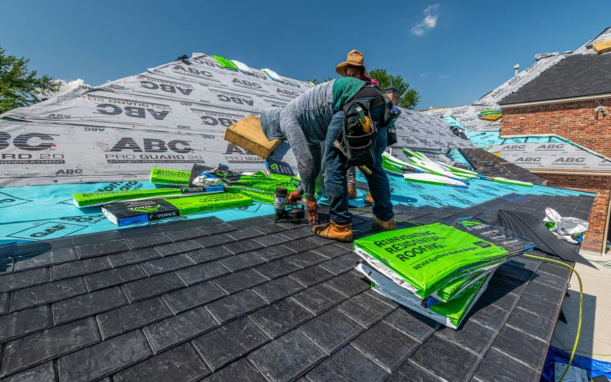 Roofer installing shingles next to bundles of roofing material on a house roof