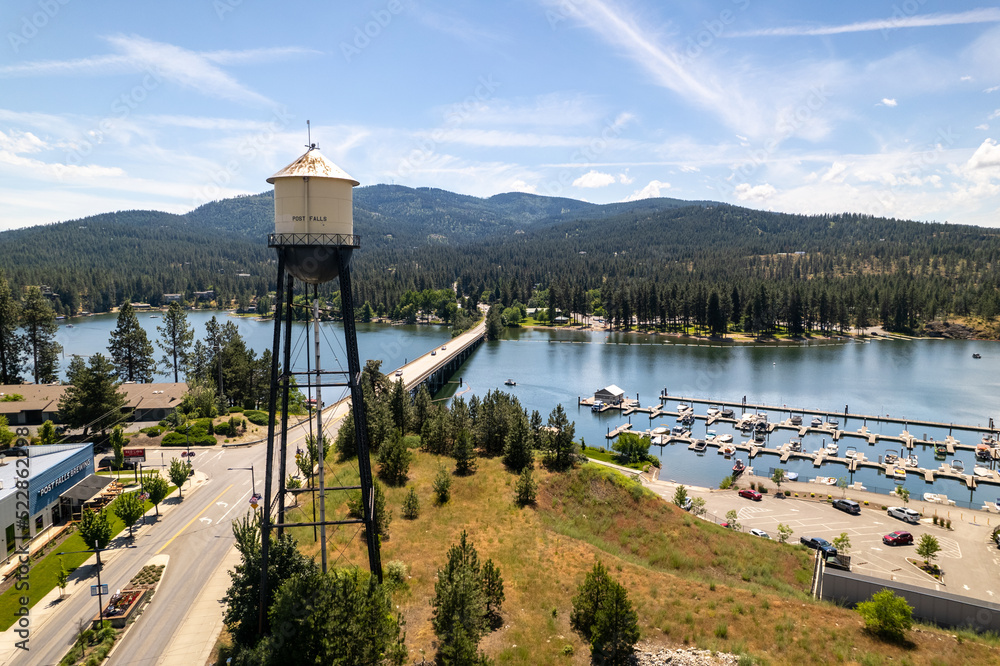 Aerial view of Post Falls, Idaho, featuring the historic 'POST FALLS' water tower, the Spokane River crossed by a long bridge, a small-boat marina, and forested rolling hills under a bright blue sky