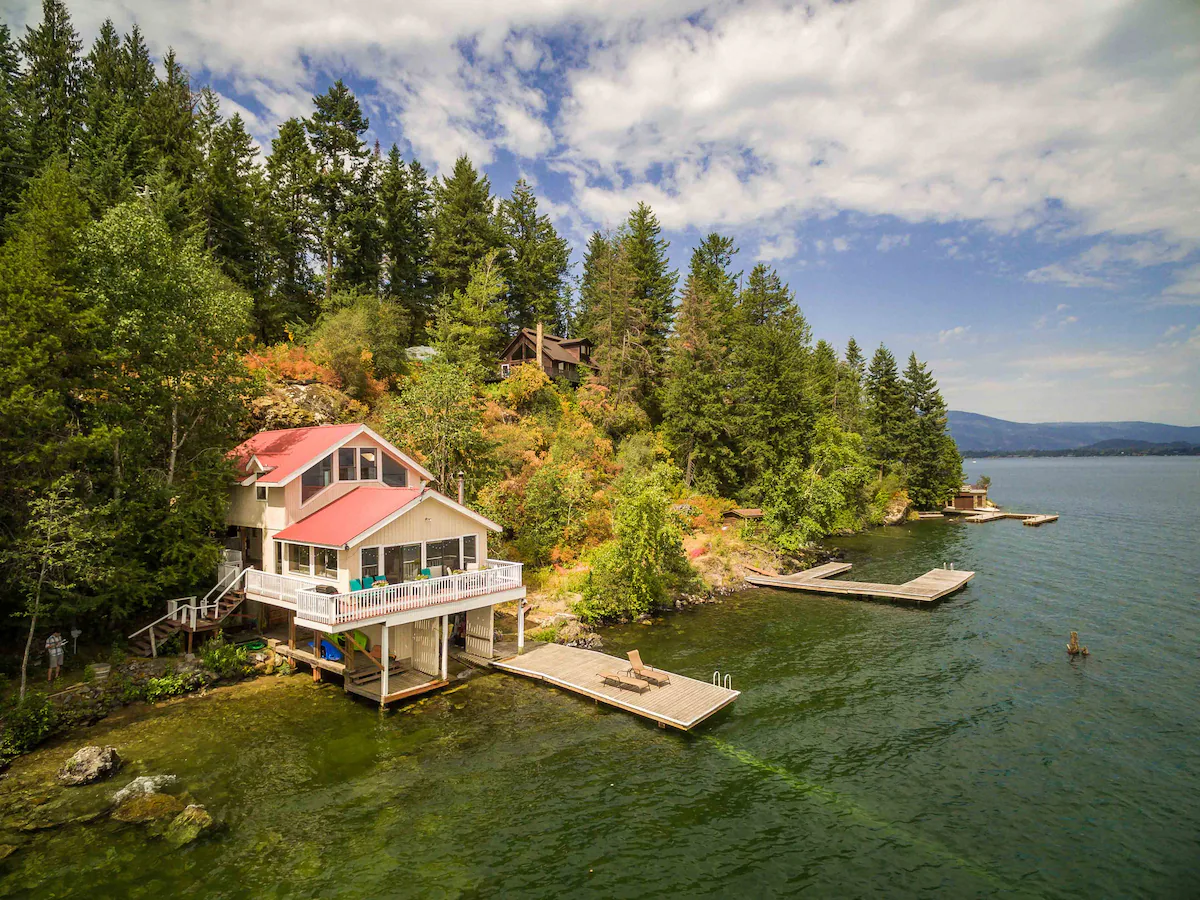 Two-story lakefront cottage with light beige siding, white trim, and a bright red metal roof, perched on pilings over a teal-green lake with wooden floating docks and a forested shoreline rising behind it