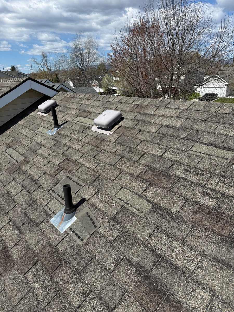 Wind-damaged asphalt shingle roof with missing shingle tabs around vents, exposing nail lines and the underlying roof decking